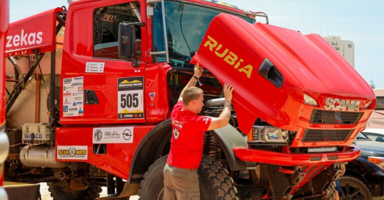 Mechanic checking a red rally truck's engine during a desert competition event.