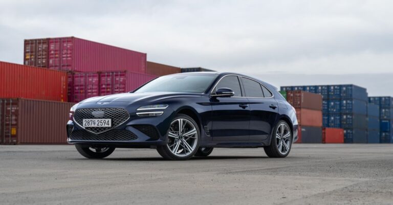 A luxury Genesis car parked in front of stacked shipping containers on a cloudy day.