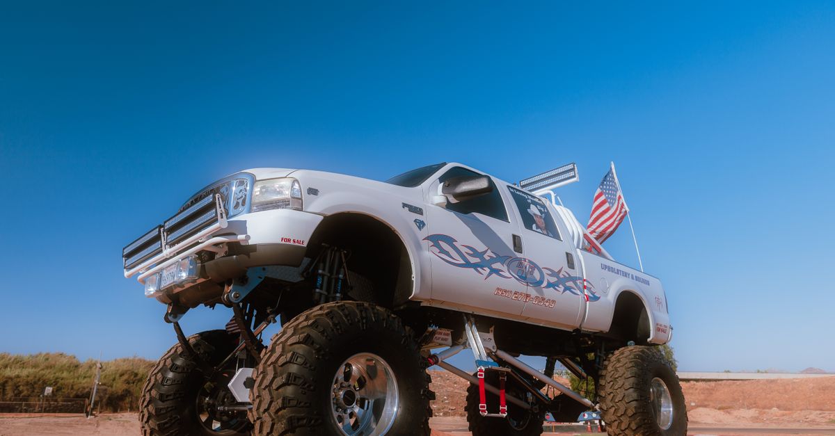 A lifted white monster truck adorned with American flags on a sunny day, showcasing off-road capability.